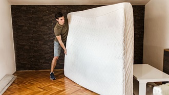 Um homem segura num colchão de casal velho que está pousado no chão por um dos lados no sentido do comprimento.